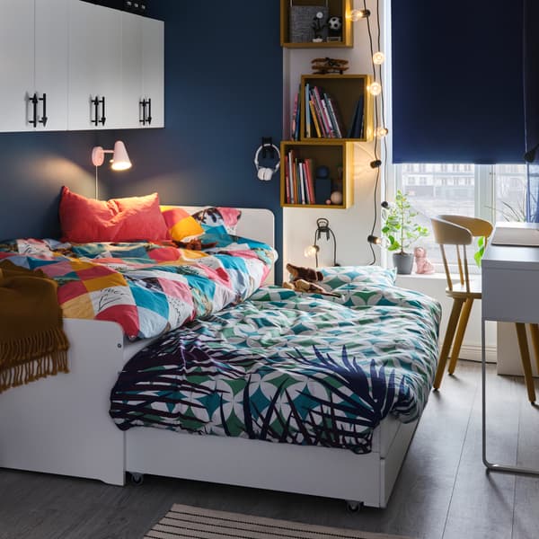 Ein weißes Bettgestell mit einem ausziehbaren Unterbett. Auf beiden Betten liegt farbenfrohe Bettwäsche. Das Rollo ist fast ganz heruntergezogen.