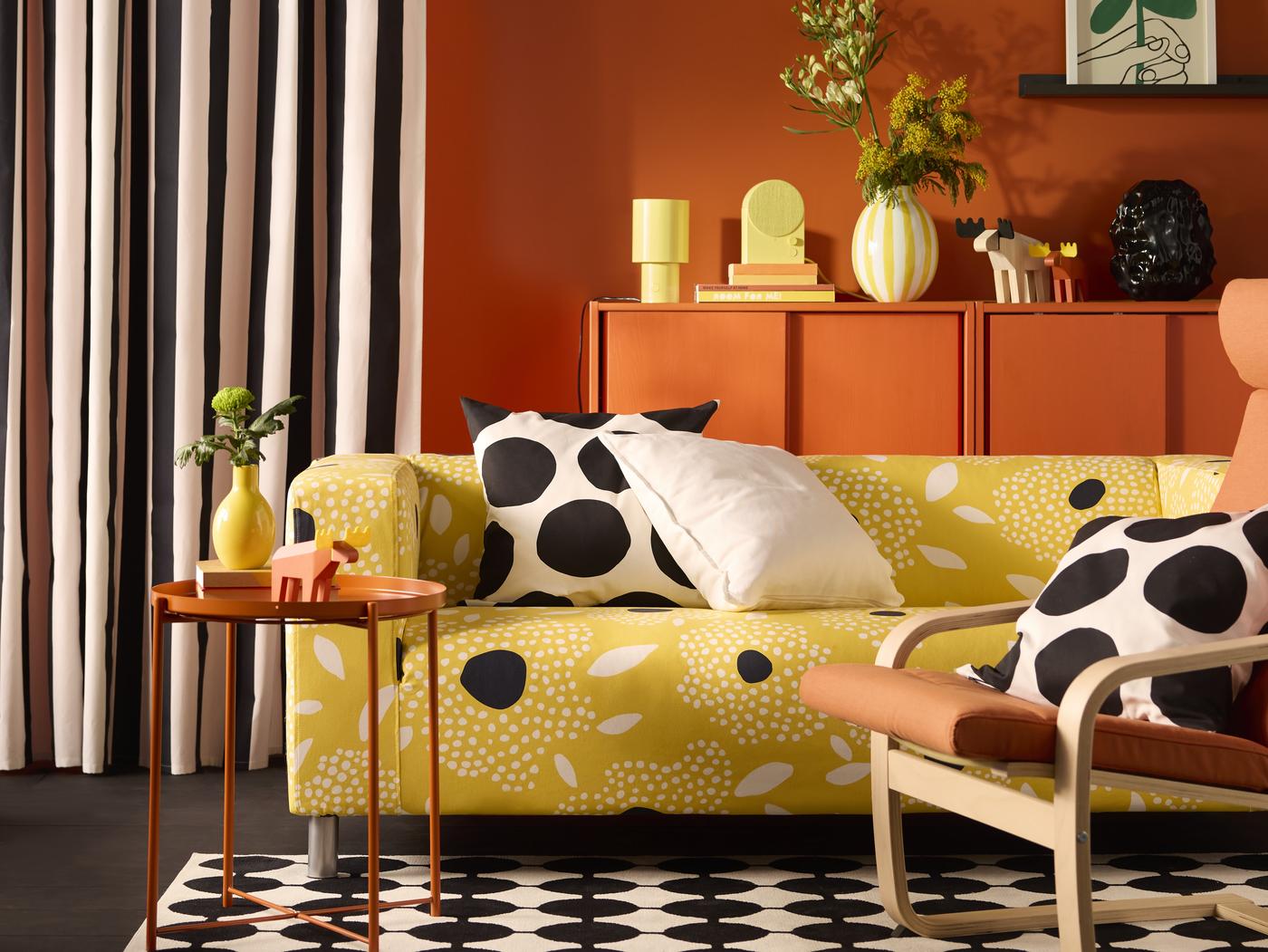 Ein Wohnzimmer im skandinavischen Stil, gelb gemustertes Sofa, schwarz-weiß getupfte Pölster und schwarz-weiß gemusterter Teppich. Im Hintergrund eine orange Kommode mit Dekorationsartikeln
