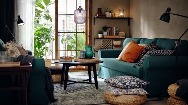 Ein Wohnzimmer mit einem dunkeltürkisfarbenen 3er-Bettsofa, einem Hocker aus Bananenstaudenfaser und einem Couchtisch.