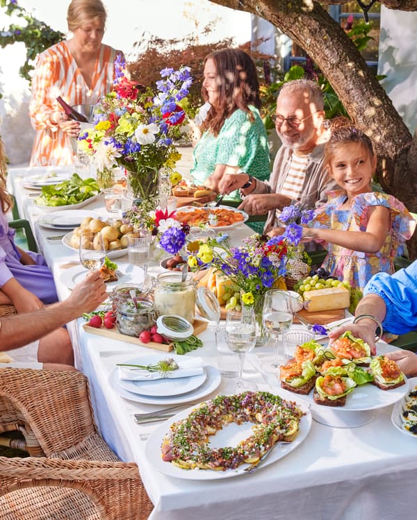 Ein wunderschön gedeckter Tisch im Freien mit weißer Tischdecke, buntem Geschirr und farbenfrohen Blumenarrangements.