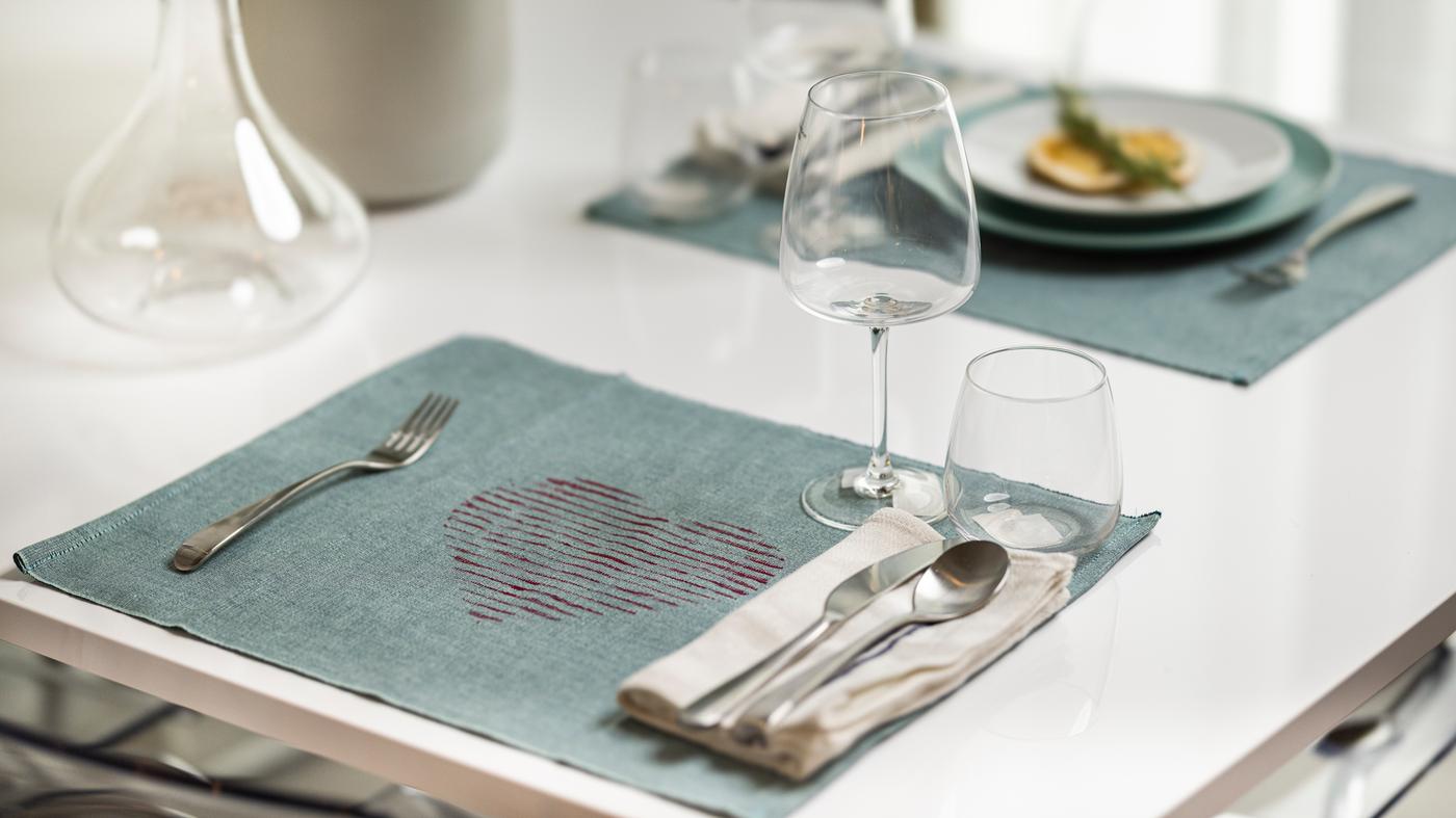 Eine blaue unterlage lirgt auf einem weißen Tisch mit einem roten Herz in der mitte. Darauf liegt Geschirr und Besteck und 1 Glas.