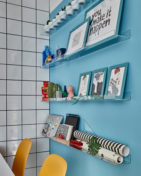 Eine blaue Wand, an der eine MELLÖSA Bilderleiste als Aufbewahrung für Bilder, Pflanzen und Geschenkpapier dient. Davor sind gelbe Stühle zu sehen.