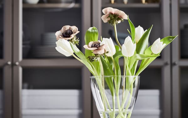 Eine durchsichtige VASEN Vase mit weissen Tulpen steht vor einer dunklen Vitrine mit Glasfronten.