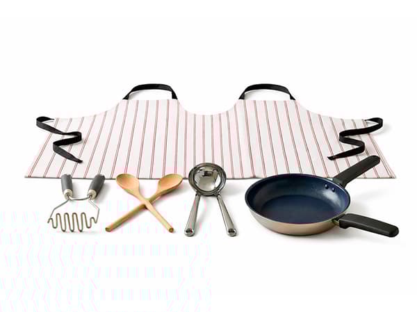 Eine fake Küchenkollektion für zwei Personen auf weißem Hintergrund. Eine breite, rot-weiß gestreifte Schürze, in der Mitte zusammengenäht für zwei Träger:innen, liegt im Hintergrund. Davor sind Küchenutensilien zu sehen: ein Kartoffelstampfer mit zwei Griffen, ein Kochlöffel für zwei, eine Schöpfkelle mit zwei Griffen aus Metall und eine blaue Bratpfanne, ebenfalls mit zwei Griffen.