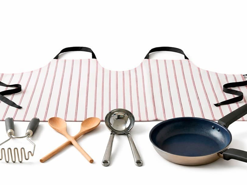 Eine fake Küchenkollektion für zwei Personen auf weißem Hintergrund. Eine breite, rot-weiß gestreifte Schürze, in der Mitte zusammengenäht für zwei Träger:innen, liegt im Hintergrund. Davor sind Küchenutensilien zu sehen: ein Kartoffelstampfer mit zwei Griffen, ein Kochlöffel für zwei, eine Schöpfkelle mit zwei Griffen aus Metall und eine blaue Bratpfanne, ebenfalls mit zwei Griffen.