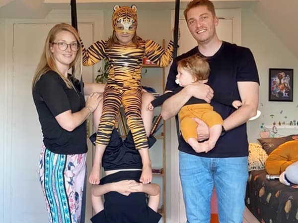 Eine Familie mit drei Kindern steht in einem Kinderzimmer. Zwei der Kids tragen Kostüme und turnen auf einer Schaukel, die an der Zimmerdecke hängt. Die Mutter hält die Schaukel stabil, der Vater hält ein Kleinkind im Arm.
