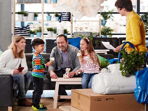 Eine Familie sitzt auf einem schwarzen Ledersofa im Eingangsbereich vom IKEA Einrichtungshaus während ein Mitarbeiter auf einem Flachwagen die Einkäufe bringt