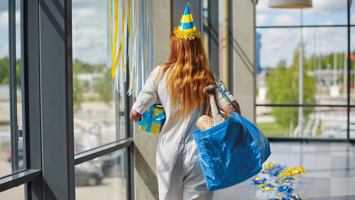 Eine Frage trägt eine Partyhüttchen auf dem Kopf, in der linken Hand ein Geschenk und auf der rechten Seite eine gefüllte IKEA FRAKTA Tasche