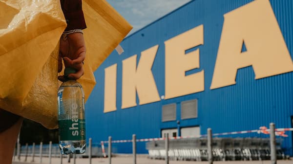 Eine Frau hält vor einem IKEA Einrichtungshaus eine share Mineralwasserflasche und eine gelde IKEA Tüte in der Hand.