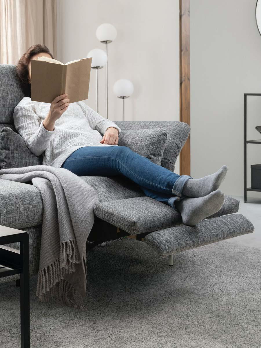 Eine Frau liegt entspannt mit einem Buch in der Hand auf einem elektrisch ausgeklappten Ruhesessel, der Teil eines hellgrauen FAMMARP Relaxsofas ist.