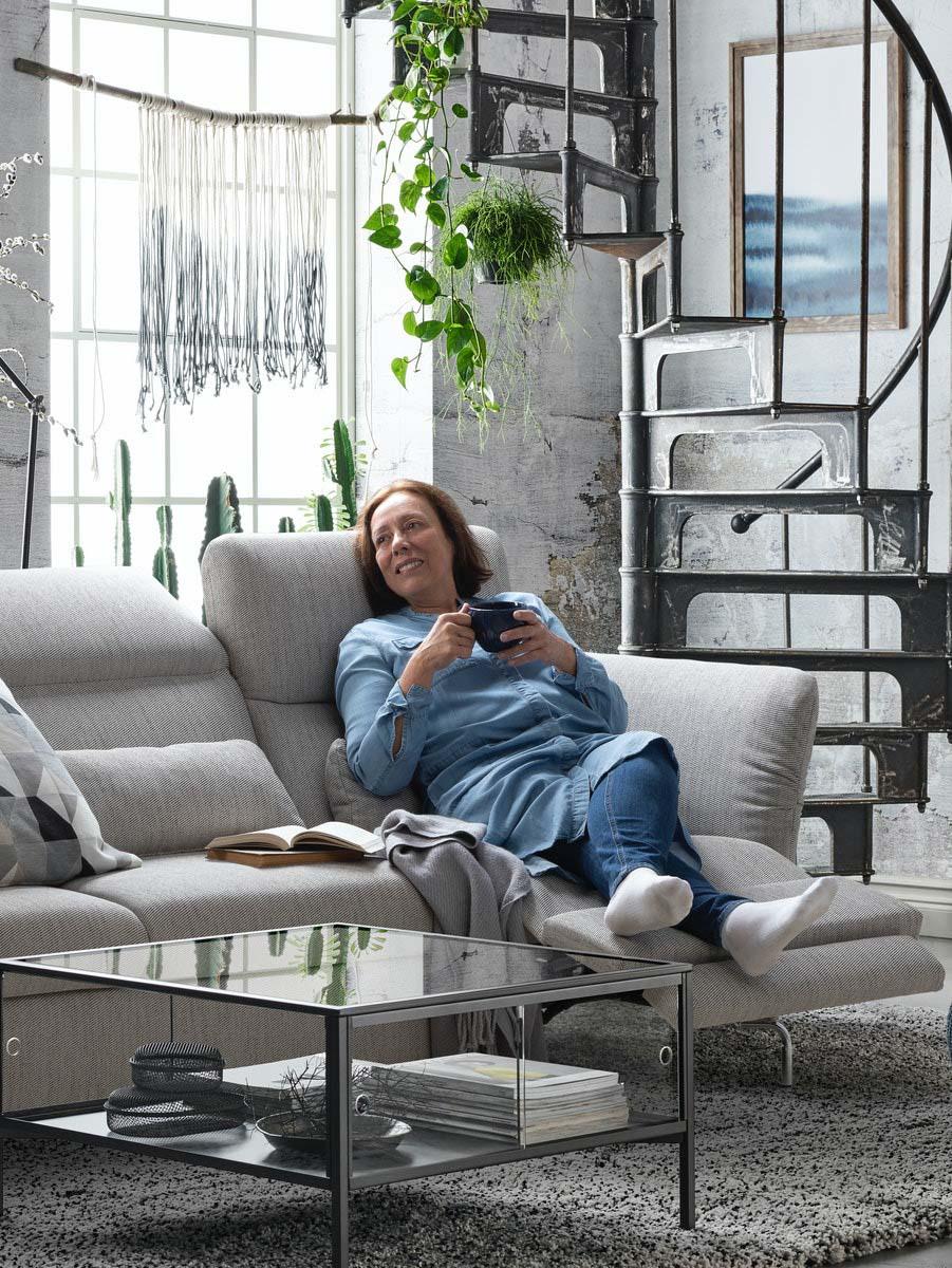 Eine Frau liegt entspannt mit einer Tasse Tee auf einem beigefarbenen Sofa mit Relaxfunktion. Die Fußstütze ist elektrisch ausgefahren.