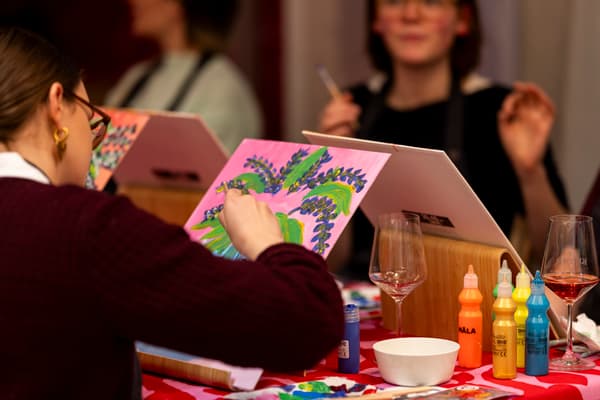 Eine Frau malt konzentriert ein buntes Bild, vor ihr stehen Farbtuben, Pinsel und ein Glas Wein.

