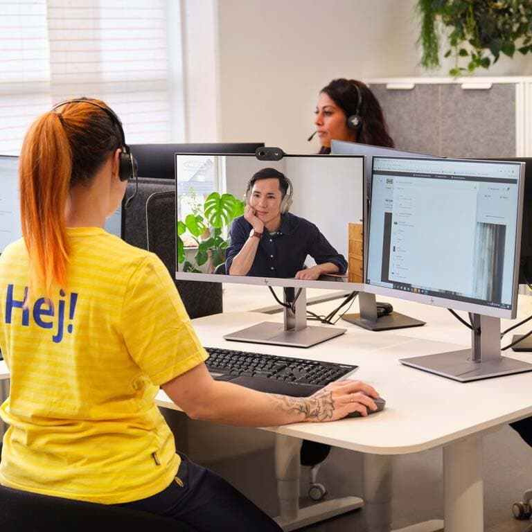 Eine Frau mit Headset und IKEA Kleidung sitzt an einem Schreibtisch vor 2 Bildschirmen. Auf dem einen Bildschirm ist ein Mann zu sehen auf dem anderen die IKEA Webseite. Die Frau führt eine Beratung per Videocall durch.