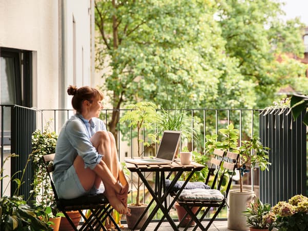 Eine Frau sitzt auf einem Balkonstuhl auf ihrem Balkon mit einem offenen Laptop auf dem Tisch.
