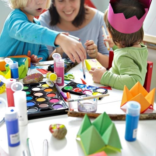 Eine Frau sitzt mit zwei Kindern am Tisch und malt. Der Tisch ist mit Papier bedeckt, u. a. zu sehen sind die MÅLA Wassermalfarben, Stifte und Farben.