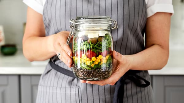 Eine Frau steht in einer Schürze in einer Küche, in den Händen ein Glas mit einem bunten Schichtsalat.