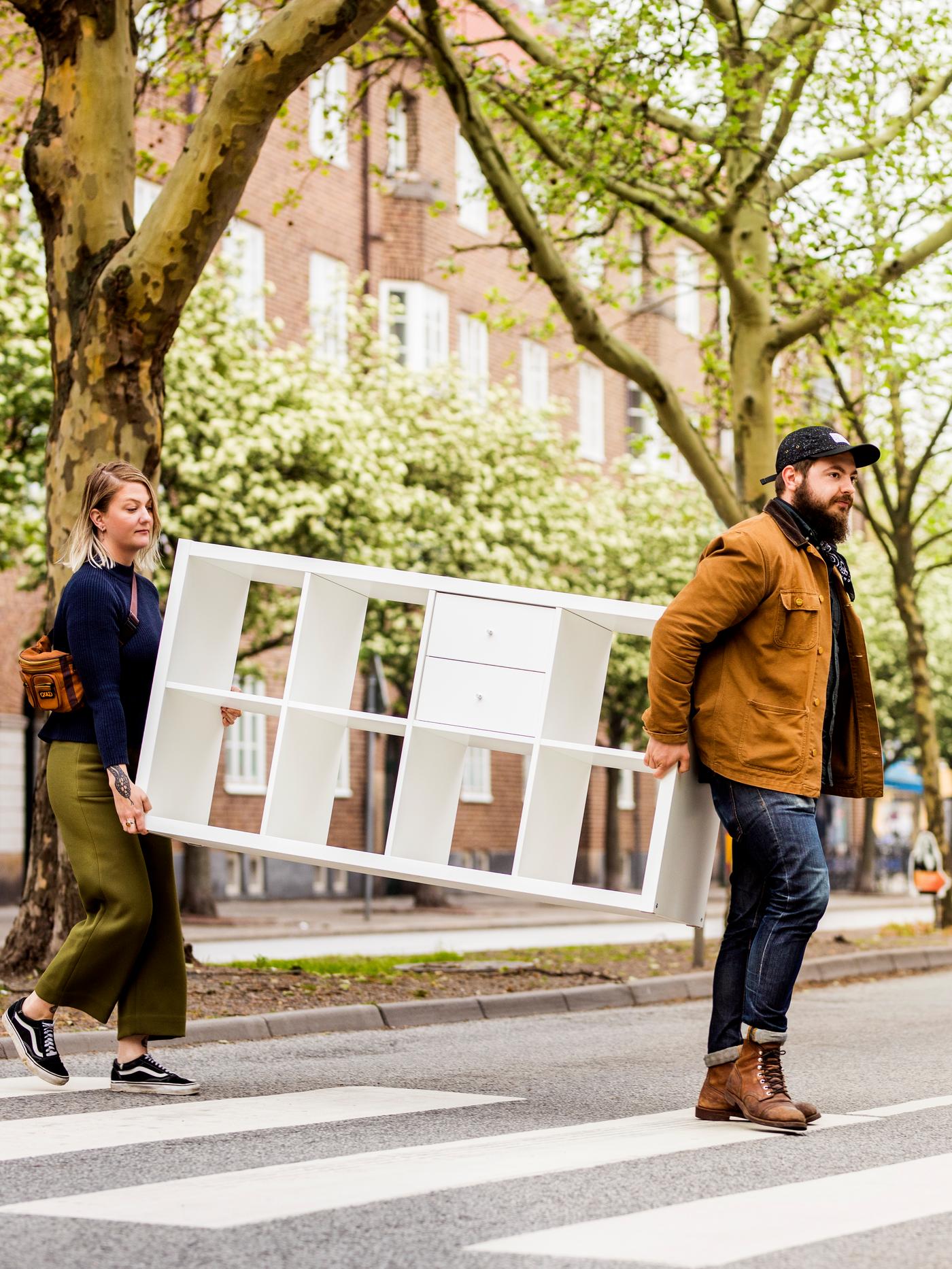 Eine Frau und ein Mann tragen ein weißes KALLAX Regal über einen Zebrastreifen