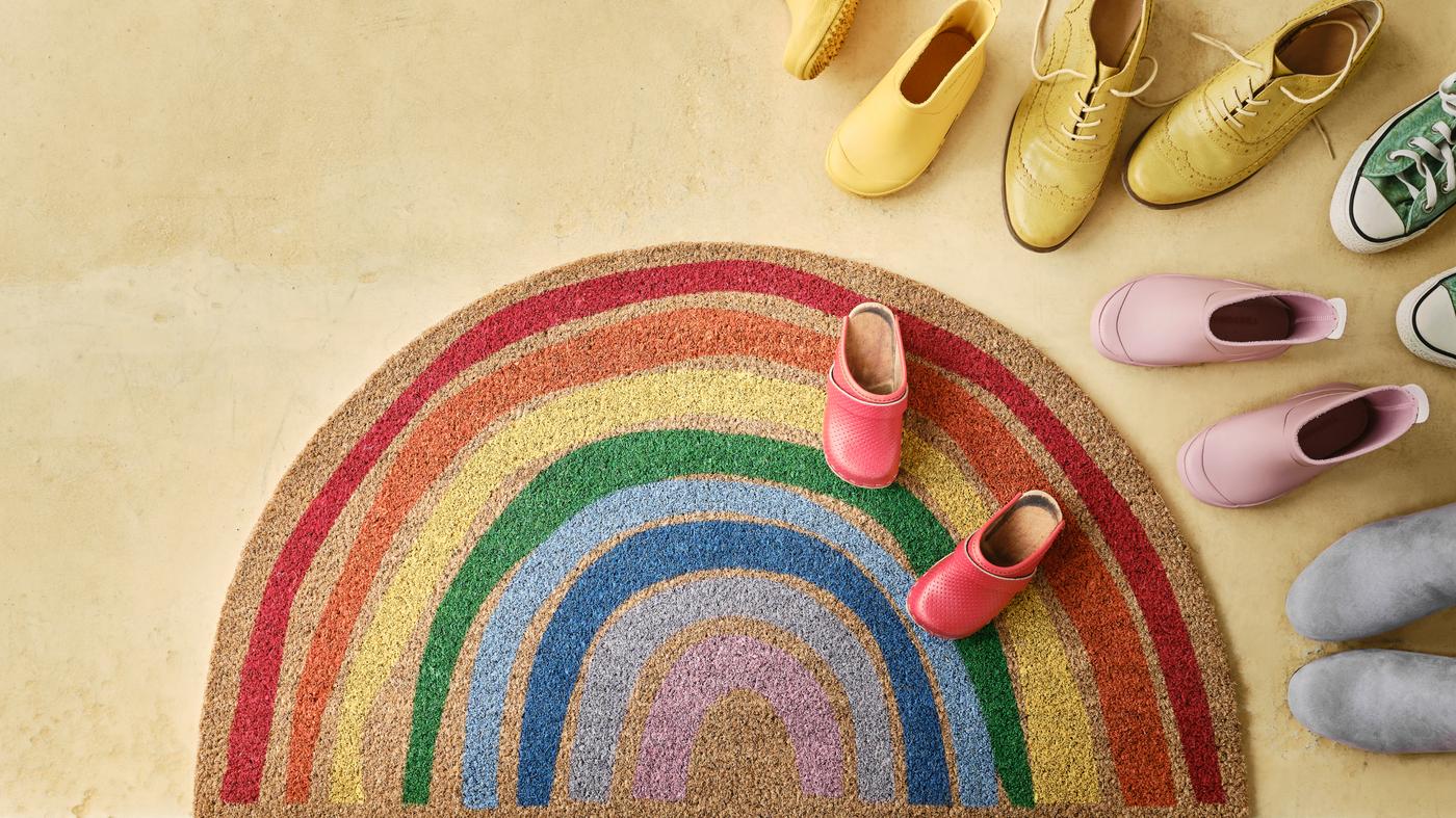Eine Fußmatte mit einem Regenbogen Motiv und auf der rechten Seite liegen verschiedene Schuhe.