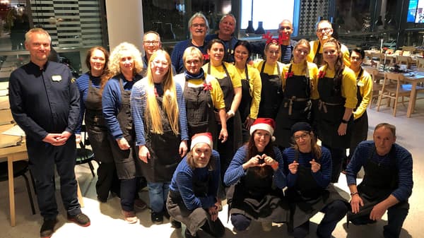 Eine grosse Gruppe von IKEA-Mitarbeitenden steht in einem festlich dekorierten Restaurantbereich. Sie tragen blaue und gelbe Arbeitskleidung mit grauen Schürzen und alle lächeln in die Kamera.