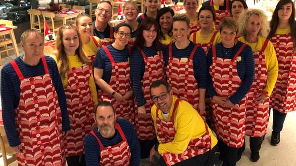 Eine grosse Gruppe von IKEA-Mitarbeitenden steht in einem festlich dekorierten Restaurantbereich. Sie tragen blaue und gelbe Arbeitskleidung mit roten Schürzen, die ein weisses Muster haben. Alle lächeln in die Kamera.