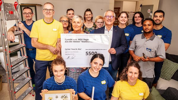 Eine große Gruppe von IKEA-Mitarbeitenden und weiteren Helfer:innen steht in einem Raum und hält einen Spendenscheck über 5.500 Euro von IKEA Regensburg für das Caritas-Projekt „Haus St. Rita“. Die Gruppe ist umgeben von Werkzeugen und einer Leiter, was auf eine Renovierungsaktion hindeutet.  