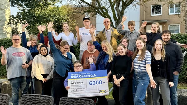Eine große Gruppe von Menschen steht draußen und winkt freudig in die Kamera. In der Mitte hält eine Person im Rollstuhl einen großen Spendenscheck von IKEA Osnabrück über 6.000 Euro für das Projekt „WirQuartier“. Die Gruppe besteht aus Personen verschiedener Altersgruppen, und im Hintergrund sind Bäume und Wohnhäuser zu sehen.  