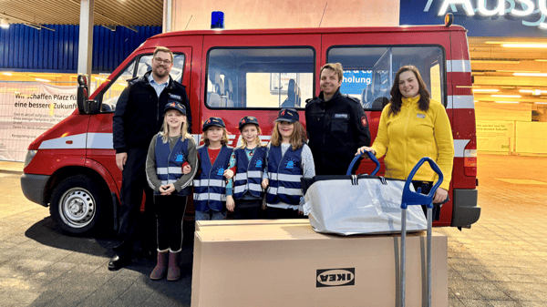 Eine Gruppe aus Feuerwehrleuten, IKEA-Mitarbeiterin und Kindern in blauen Feuerwehr-Westen steht vor einem roten Feuerwehrfahrzeug. Vor ihnen sind IKEA-Kartons und eine IKEA-Tasche auf Transportrollern gestapelt. Die Szene spielt sich vor einem IKEA-Gebäude ab.