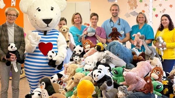 Eine Gruppe aus Krankenhauspersonal, einer IKEA-Mitarbeiterin und einer weiteren Frau steht in einem bunt dekorierten Raum mit einem grossen Maskottchen in Bärenform. Vor ihnen liegt ein grosser Berg aus Kuscheltieren, die sie für eine Spendenaktion übergeben.