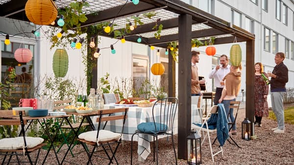 Eine Gruppe Menschen steht neben einer Reihe von Stühlen und Tischen unter einer Pergola in einem Innenhof. Auf dem Tisch stehen leckere Snacks.