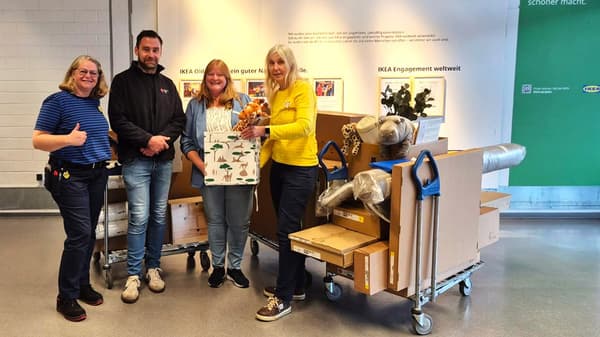 Eine Gruppe von 4 Personen steht in einem IKEA-Geschäft vor einer Wand mit dem Schriftzug „Gemeinschaften unterstützen, wo immer wir sind“. Sie haben Einkaufswagen voller Möbel, Decken und Haushaltswaren und zeigen mit Daumen nach oben.