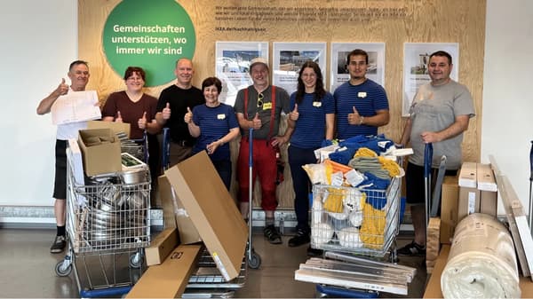 Eine Gruppe von acht Personen steht in einem IKEA-Geschäft vor einer Wand mit dem Schriftzug „Gemeinschaften unterstützen, wo immer wir sind“. Sie haben Einkaufswagen voller Möbel, Decken und Haushaltswaren und zeigen mit Daumen nach oben.