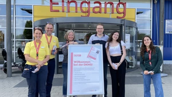 Eine Gruppe von IKEA-Mitarbeitenden, eine DKMS Helferin und weiteren Personen stehen lächelnd gemeinsam mit einem DKMS Info-Schild vor dem IKEA Braunschweig Eingang.