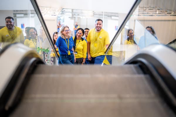 Eine Gruppe von IKEA-Mitarbeitern in gelben und blauen Uniformen steht in einer hellen, modernen Umgebung neben einer Rolltreppe.
