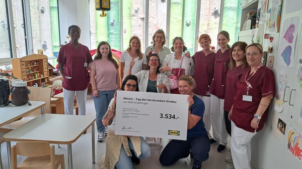 Eine Gruppe von medizinischem Personal in bordeauxroten Kitteln, einer Clown-Doktorin und weiteren Unterstützer:innen steht in einem kinderfreundlich gestalteten Raum. Zwei Personen knien vorne und halten einen Spendenscheck über 3.534 Euro von IKEA Sindelfingen für die Olgäle-Stiftung für das kranke Kind e. V. im Rahmen der Aktion „Tag des herzkranken Kindes“.  