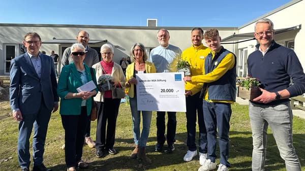 Eine Gruppe von neun Personen stehen auf einer Wiesenfläche. Unter anderemzwei IKEA Mitarbeitende und die Leiterin des Kindergartens posieren mit einem 20.000€ Spendenscheck. Im Hintergrund lder Kindergarten, der renoviert wurde.