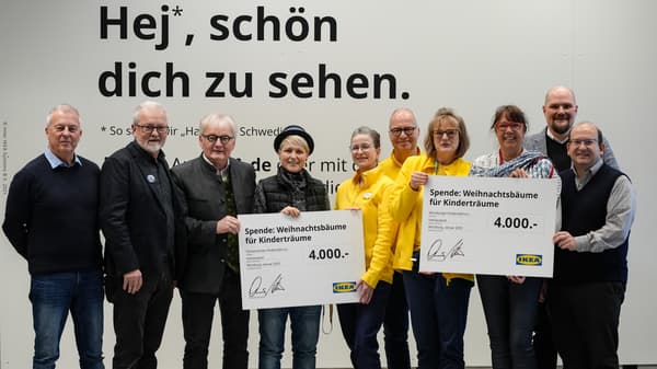 Eine Gruppe von neun Personen steht vor einer weissen Wand mit der IKEA-Aufschrift „Hej, schön dich zu sehen.“ Drei der Personen tragen gelbe IKEA-Dienstkleidung. Zwei Personen halten grosse Spendenschecks in Höhe von jeweils 4.000 Euro mit der Aufschrift „Spende: Weihnachtsbäume für Kinderträume“. Die Gruppe lächelt in die Kamera.  
