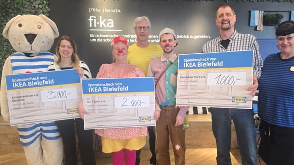 Eine Gruppe von sechs Personen, darunter zwei als Clowns und eine in einem Bärenkostüm, hält drei große Spendenschecks von IKEA Bielefeld in den Händen. Die Schecks sind für „Kinderzentrum e.V.“, „Dr. Clown e.V.“ und „Bielefelder Tisch e.V.“ ausgestellt