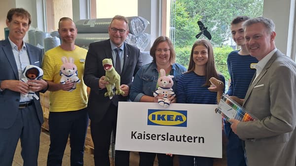 Eine Gruppe von sieben Personen, darunter IKEA-Mitarbeitende und weitere Beteiligte, posiert gemeinsam mit Kuscheltieren und einem IKEA-Kaiserslautern-Schild. Sie stehen in einem Raum mit gestapelten Matratzen im Hintergrund. Einige halten Plüschtiere, während eine Person eine Packung Filzstifte präsentiert.