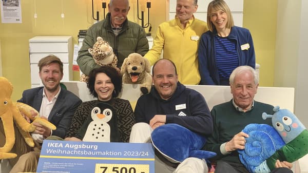 Eine Gruppe von sieben Personen sitzt und steht in einem IKEA-Möbelhaus, umgeben von Kuscheltieren. Sie halten ein Schild mit der Aufschrift „IKEA Augsburg Weihnachtsbaumaktion 2023/24“ und der Spendensumme 7'500 Euro. Alle lächeln in die Kamera.