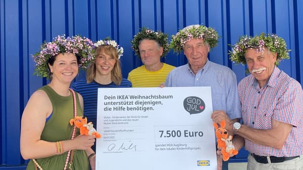 Eine Gruppe von vier Personen mit Blumenkränzen auf dem Kopf, steht vor dem Einrichtungshaus. Sie halten ein Schild mit der Aufschrift „IKEA Augsburg Weihnachtsbaumaktion 2024/25“ und der Spendensumme 7'500 Euro. Alle lächeln in die Kamera.