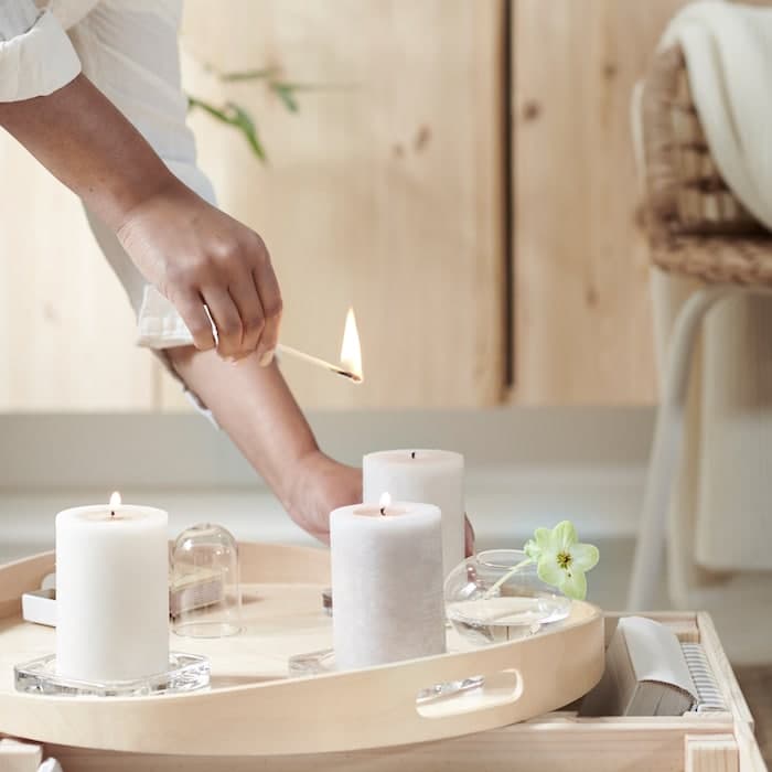 Eine Hand zündet drei LUGGA Duftkerzen an, die auf einem Holztablett vor einem IVAR Schrank stehen.