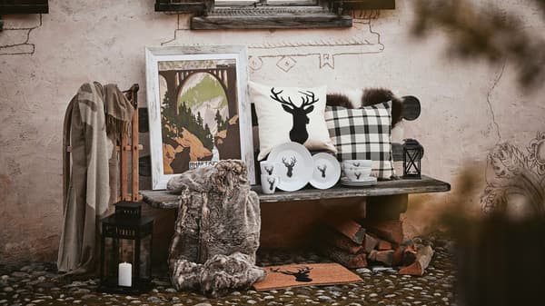 Eine Holzbank vor einer Wand eines alten Chalet, auf der viele Produkte der IKEA Chalet-Kollektion stehen, wie z.B. Kissen, ein gerahmtes Bild, flauschige Decken, Geschirr mit Hirschmotiv und ein Fell.