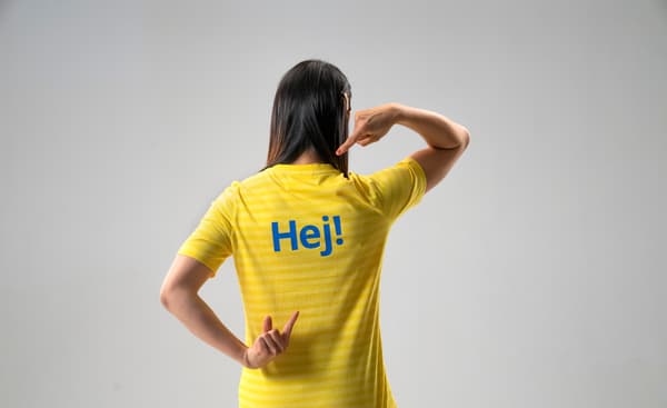 Eine IKEA Mitarbeiterin im gelben T-Shirt steht mit dem Rücken zugewandt und zeigt auf das "hej!", welches auf ihrem Rücken steht 