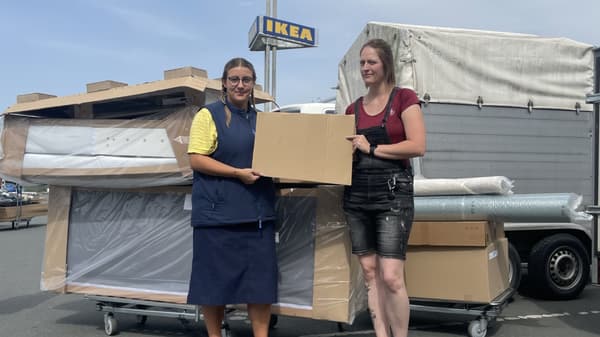 Eine IKEA-Mitarbeiterin in blauer und gelber Kleidung und eine weitere Frau stehen draussen auf einem Parkplatz vor einem grossen Stapel Möbelkartons. Sie halten gemeinsam ein Pappschild mit der Aufschrift „Spende“. Im Hintergrund ist das IKEA-Logo auf einem Schild zu sehen.