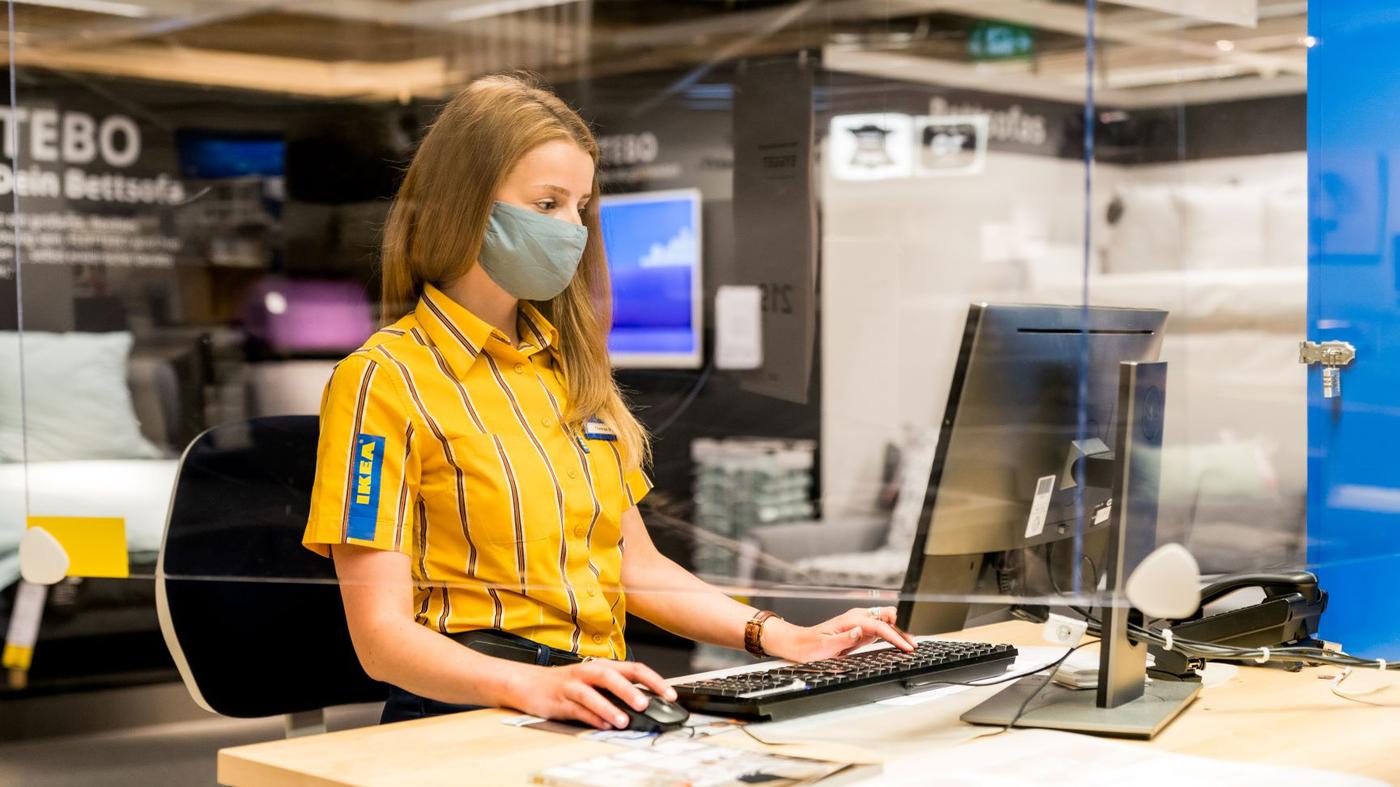 Eine IKEA Mitarbeiterin sitzt vor einem Computer, sie trägt ein gelbes Hemd und eine hellblaue Maske.