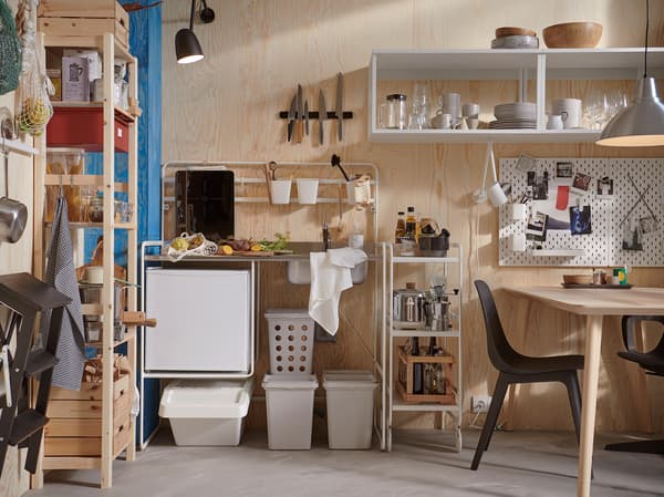 Eine kleine Küche mit einer SUNNERSTA Miniküche, grauen Recyclingbehältern, schwarzen Stühlen, Kühlschrank, Regal und Hängeleuchte.
