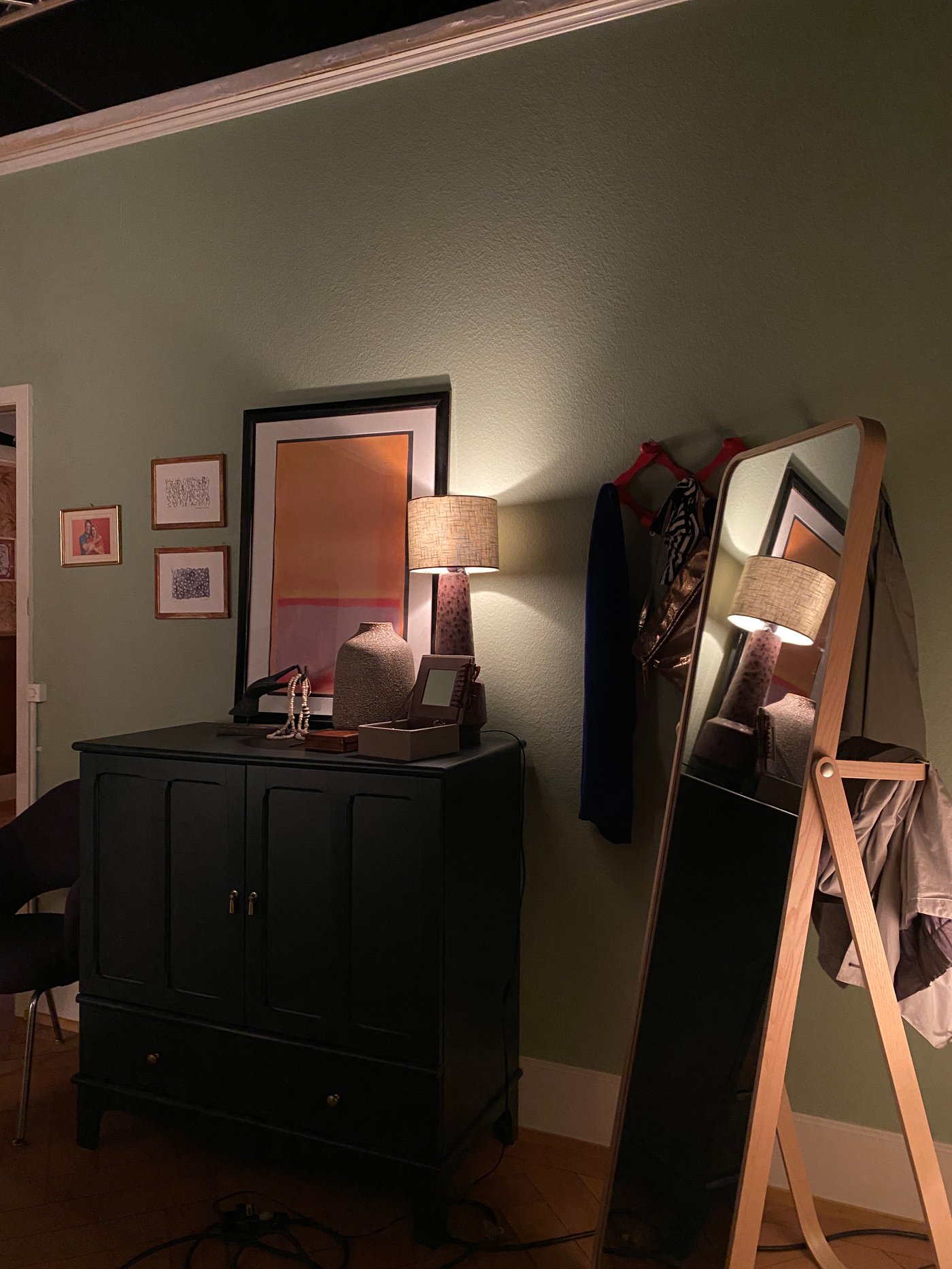 Eine Kommode in einem Schlafzoimmern mit Bildern und einer Tischlamoe darauf neben einem Standspiegel und Garderobenhaken an der Wand.