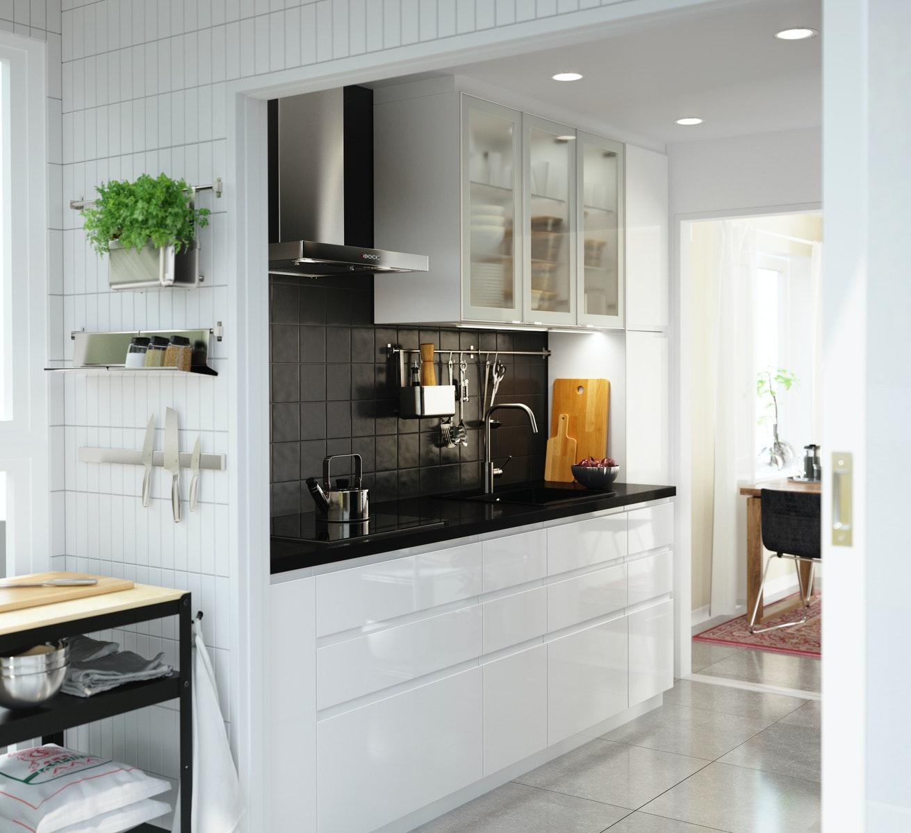 Eine Küche mit den VOXTORP Fronten in Hochglanz Weiß mit schwarz gefliester Wand, schwarzer Arbeitspatte und einer Stange, an der Küchenutensilien hängen