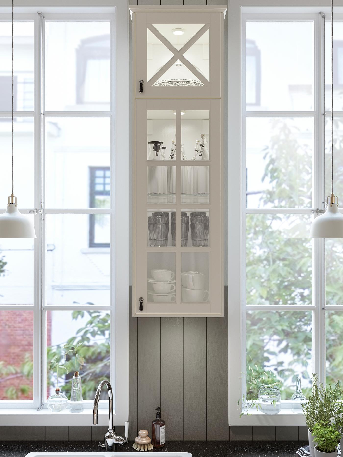 Eine Küche mit einem schmalen Wandschrank mit BODBYN Vitrinentüren zwischen zwei Fenstern. Die Arbeitsplatte ist in Schwarz gehalten.