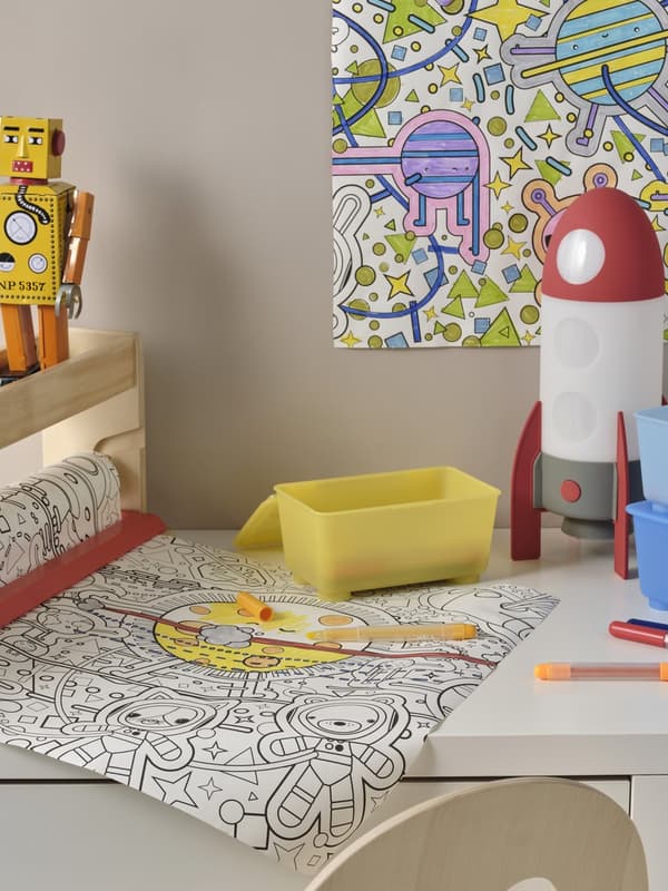 Eine Malpapierrolle mit Weltraum Figuren zum Ausmalen liegt mit Stiften auf einem Tisch. 
